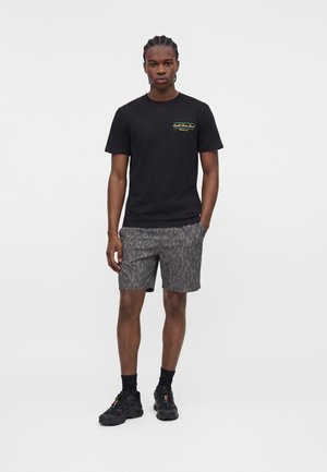 Giovane uomo in piedi che indossa una t-shirt nera con grafica, pantaloncini grigi con motivo, calzini neri e sneakers nere su sfondo uniforme.