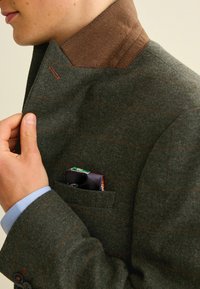 Dunkelgrüner Wollblazer mit braunem Kragen, orangefarbenen Nadelstreifen und einem Einstecktuch, das aus der Brusttasche hervorlugt. Texturierte Stoffdetails sichtbar.