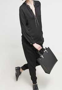 Personne portant un haut à manches longues noir et un pantalon, tenant un grand sac fourre-tout noir avec des accents métalliques, debout avec une jambe légèrement levée.