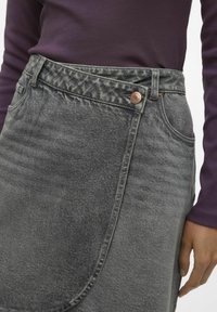 Falda de mezclilla gris con cierre de botón, textura sutil y panel frontal en ángulo. Combinada con una blusa ajustada de manga larga morada.