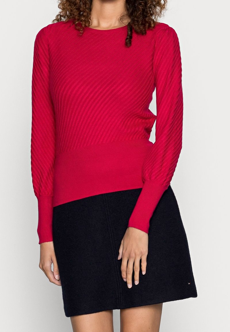 Femme portant un pull rouge ajusté à manches longues avec des côtes diagonales et une jupe noire évasée, debout devant un fond uni.