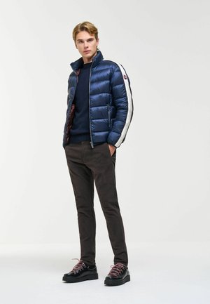 Giovane uomo in piedi che indossa una giacca imbottita blu navy con strisce bianche, pantaloni scuri, scarpe nere con lacci rossi, mani nelle tasche.