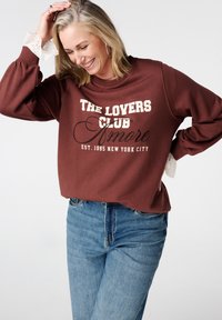 Bruine trui met "THE LOVERS CLUB Amore" gedrukt in crème en zwart. Heeft bolvormige mouwen met kanten afwerkingen. Draagt bij lichte denim jeans.