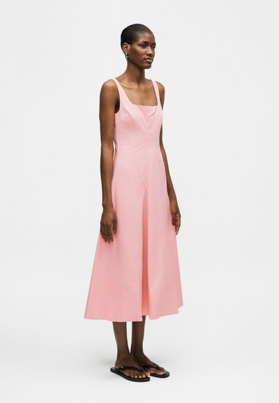 Γυναίκα όρθια, φοράει ροζ φορέμα μίντι χωρίς μανίκια με τετράγωνο ντεκολτέ και μαύρα επίπεδα σανδάλια, σε απλό ανοιχτό φόντο.