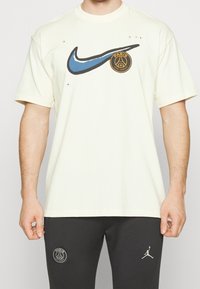 Kremowa koszulka z krótkim rękawem z niebieskim logo Nike w kształcie 'swoosh' oraz czarnym logo Paris Saint-Germain na lewej piersi. Luźny krój.