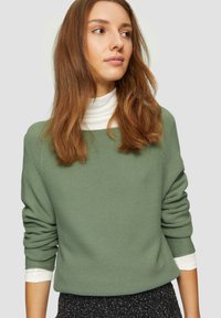 Grüner, strukturierten Pullover mit weitem Ausschnitt und langen Ärmeln, über einem weißen Rollkragenpullover getragen. Ein dunkles gemustertes Gewebe ist teilweise sichtbar darunter.