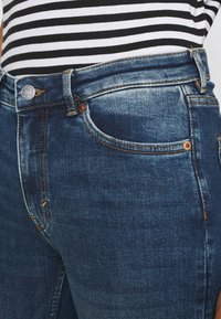 Jeans de mezclilla azul oscuro con costuras en contraste, que cuentan con un bolsillo frontal, un cierre de botón metálico y un diseño de corte ajustado.