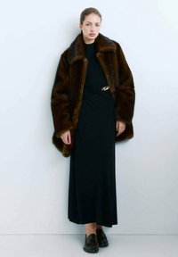 Manteau en fausse fourrure marron avec un large col, porté sur une robe noire à manches longues ornée d'une broche dorée, associé à des mocassins noirs.