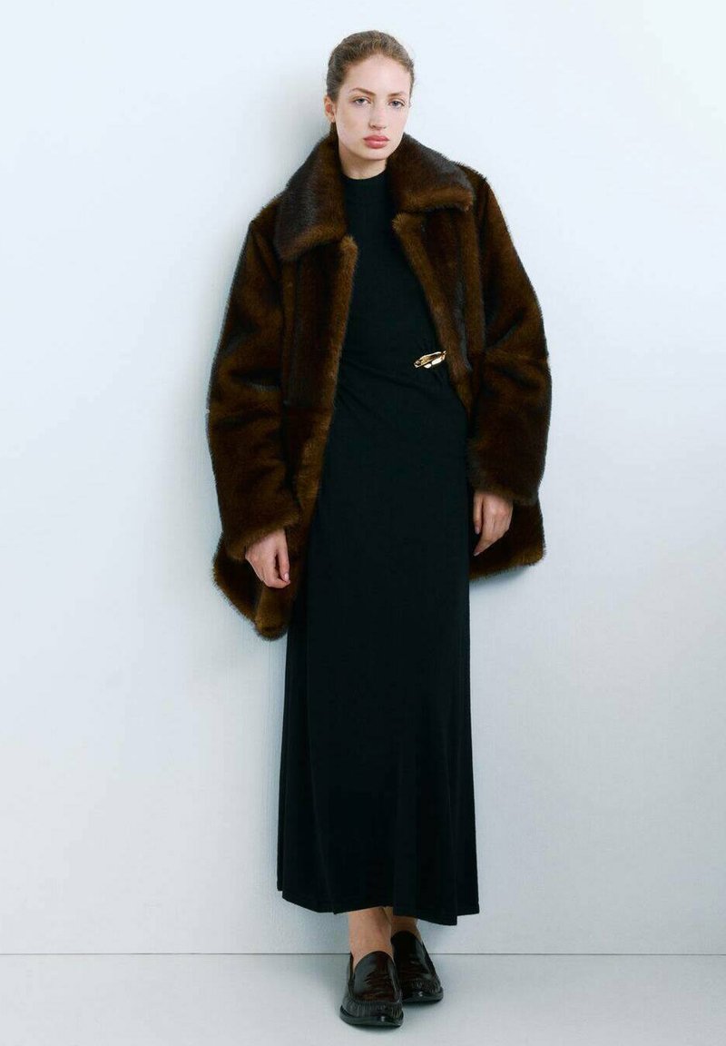 Manteau en fausse fourrure marron avec un large col, porté sur une robe noire à manches longues ornée d'une broche dorée, associé à des mocassins noirs.