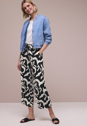 Giovane donna con capelli corti biondi indossa una giacca bomber azzurro chiaro, top bianco, pantaloni larghi a fantasia bianco e nero e sandali neri senza lacci.