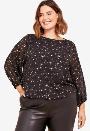 Schwarze, durchsichtige Bluse mit silbernen Sternmustern, runder Ausschnitt und lockeren Ärmeln; kombiniert mit schwarzen, lederähnlichen Hosen.