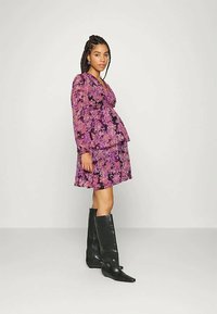 Mulher vestindo um vestido floral roxo de mangas longas e botas pretas até ao joelho, de pé de lado contra um fundo simples.
