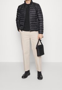 Schwarze Steppjacke mit Stehkragen, kombiniert mit beigen Hosen. Schwarze Chelsea-Stiefel und eine kleine schwarze Handtasche.