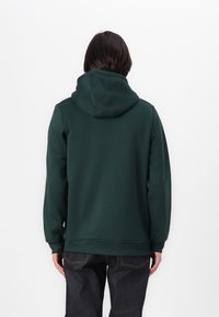 Sötétzöld kapucnis pulóver, húzózsinórral, puha anyagból készült. Részletes bordázott mandzsettákkal és aljával rendelkezik. Sötét farmernadrággal viselve, hátulról nézve.