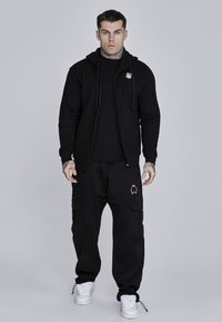 Črna zip-up hoodie s majhnim logotipom, v kombinaciji s širokimi črnimi cargo hlačami, ki imajo stranske žepke in zlato akcentno podrobnost.