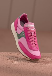 Rozā kedas ar zamšādu un sintētisku materiālu, iekļaujot zaļus akcentus, apaļas šņores un baltu zolīti. Saucony logo uz mēles.