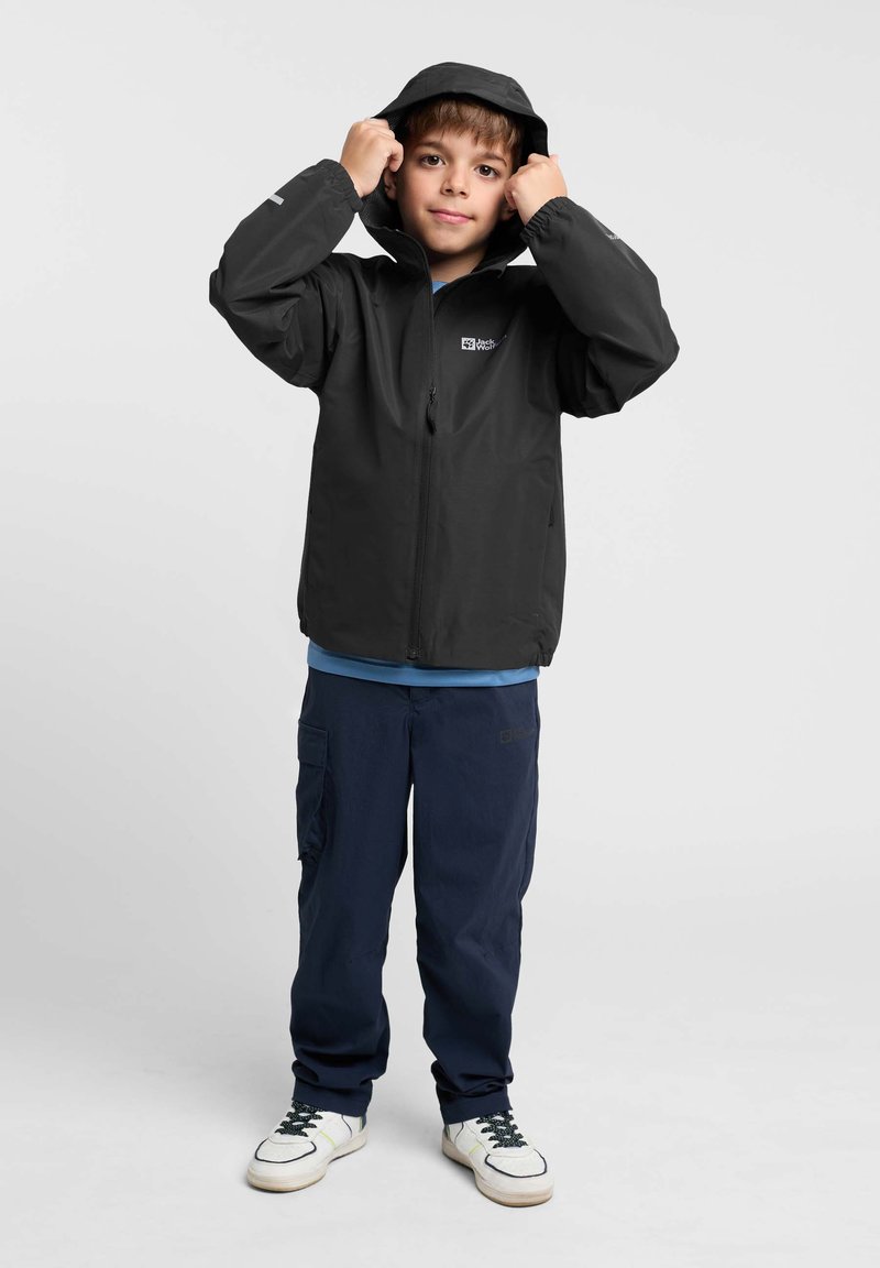Zwarte met rits gesloten jas met capuchon, gedragen over een blauwe shirt. De jongen draagt donkere broek en sneakers, staand tegen een lichte achtergrond.