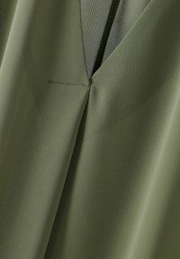 Olivengrüner Stoff mit strukturierter Oberfläche. Verfügt über ein V-Ausschnitt-Design und eine sichtbare Naht. Das Material wirkt leicht und glatt.
