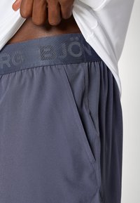 Ανδρικές αθλητικές βερμούδες με σκούρο γκρι ύφασμα, ελαστική μέση με εγχάρακτο λογότυπο και πλαϊνές τσέπες. Λείος υφς. συνολικά.