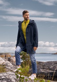 Homme portant un imperméable sombre par-dessus une veste jaune et un jean, debout sur des rochers près de l'eau sous un ciel nuageux.