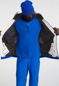 Blauwe jas, ingezip met een bijpassende blauwe lange mouw top eronder. Heeft een ritssluiting bij de hals, een soepele textuur en binnenzakken.