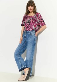 Top con stampa floreale in tonalità di rosa e viola, abbinato a jeans azzurri chiari a vestibilità comoda. Scarpe beige con suole nere texturizzate.