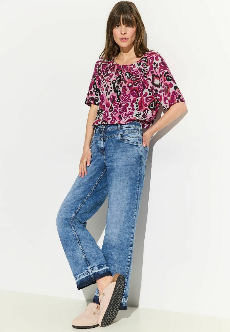 Top con stampa floreale in tonalità di rosa e viola, abbinato a jeans azzurri chiari a vestibilità comoda. Scarpe beige con suole nere texturizzate.