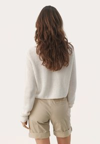 Suéter de punto en crema, con un corte holgado y textura acanalada, combinado con pantalones cortos beige enrollados. Vista trasera, diseño minimalista, fondo liso.