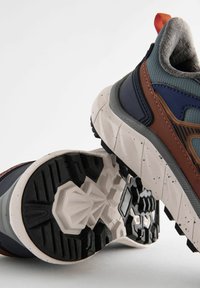 Chaussures de sport avec une tige en mesh gris, des accents marron et marine, et une semelle en caoutchouc noir texturée avec un motif de bande de roulement multidirectionnel.