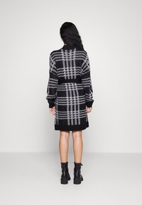 Robe à carreaux noir et blanc, longueur genoux, à manches longues, en tissu tricoté texturé, avec un col rond. Des bottines noires à la cheville complètent le look.