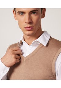 Beżowa kamizelka swetrowa z dekoltem w szpic, wykonana z dzianiny, noszona na białej koszuli z kołnierzykiem, z teksturowanym wykończeniem i gładkim dekoltem.