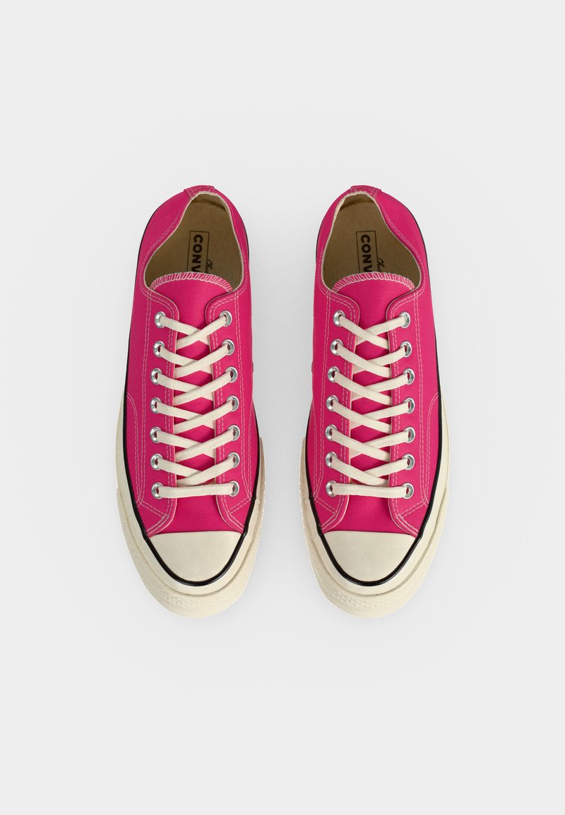 Helle pinke Low-Top-Canvas-Sneakers mit weißen Gummisohlen und cremefarbenen Schnürsenkeln, von oben auf hellem Hintergrund betrachtet.