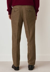 Brown houndstooth trousers with a straight cut, featuring two back pockets and black buttons, paired with black shoes, viewed from the back.