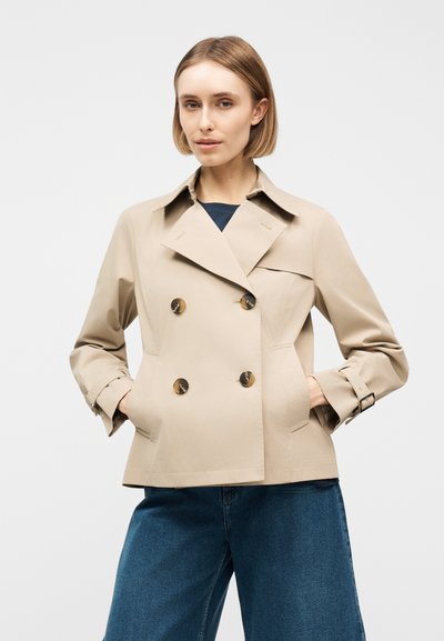 Femme aux cheveux blonds courts portant un trench-coat beige croisé et un jean bleu à jambes larges, sur un fond clair uni.