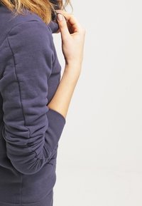 Mörkblå sweatshirt med ribbade muddar och en avslappnad passform, med hög krage och mjukt bomullsmaterial, som visar en lätt struktur.