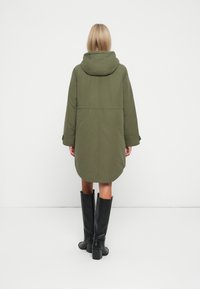 Lang olivengrøn parka med hætte, der har en afslappet pasform og detaljer i bagsømmen. Kombineret med knæhøje sorte støvler med hæl.