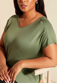 Grön t-shirt med kort ärm och V-ringning, gjord av mjukt tyg, med en avslappnad passform och lätt glans, som är kombinerad med mörka underdelar och händerna vilande på en stol.
