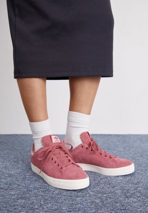 Ben iført hvide ribstrikkede sokker og lyserøde Adidas ruskinds-sneakers, stående på blåt tæppe, med nederdelens mørke kant synlig over læggene.