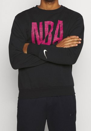 Person trägt schwarzen Pullover mit großem pinkfarbenem "NBA"-Schriftzug und weißem Nike-Swoosh auf dem Ärmel, Arme vor der Brust verschränkt.