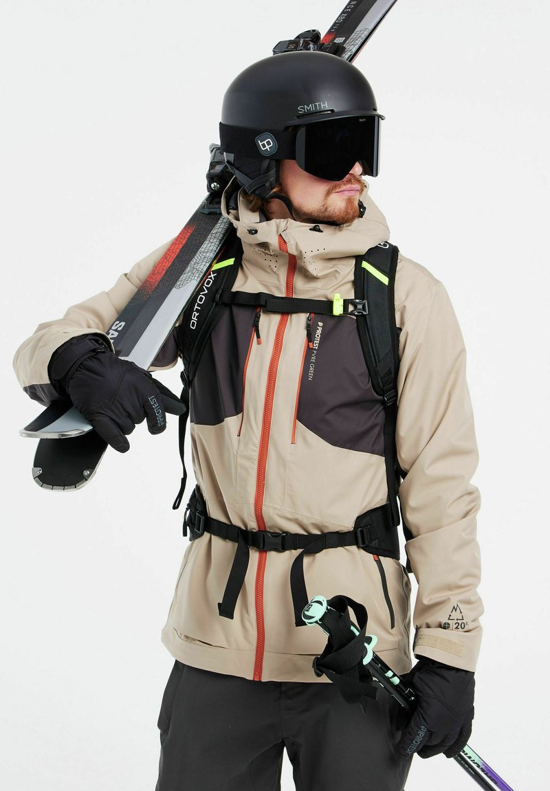 Ski-jack in beige en zwart met oranje ritsen, uitgerust met een zwarte rugzak en handschoenen, terwijl hij skies en skistokken vasthoudt.