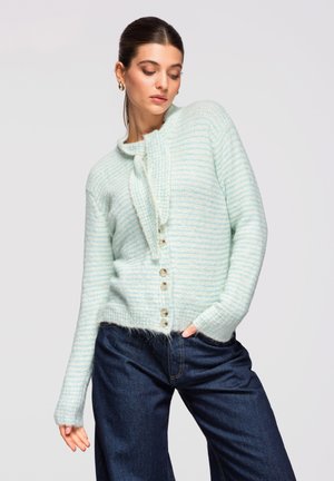 Donna che indossa un cardigan a righe azzurro chiaro e bianco con bottoni e cravattino al collo, abbinato a jeans blu scuro, in posa con una mano in tasca.