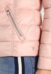 Jasnoróżowa pikowana kurtka z zamkiem błyskawicznym i kieszenią, gładka tekstura, poziome pikowanie oraz srebrne akcenty.