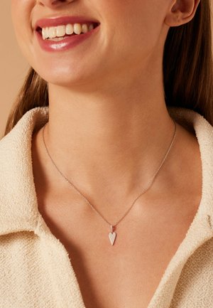 Smiling person wearing a beige textured collar garment and a silver chain necklace with a small arrowhead-shaped pendant.