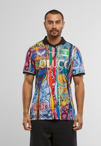 Bărbat cu păr scurt și barbă, îmbrăcat cu o căma�șă cu un model abstract multicolor, cu guler negru și pantaloni scurți negri, stând în fața unui fundal alb.