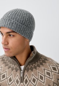 Gorro de punto gris acanalado de lana, diseño ajustado. Combina con un suéter marrón claro con patrones de diamante y cuello con cremallera.