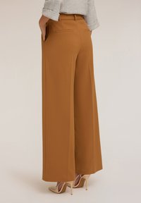 Un pantalon large dans une teinte marron chaud, fabriqué en tissu lisse. Il dispose de deux poches arrière et d'une ceinture assortie, associé à des talons de couleur claire.