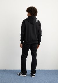 Felpa nera con cappuccio e logo sulla schiena, abbinata a jeans neri e sneakers bianche e nere. Ambientata su uno sfondo chiaro.