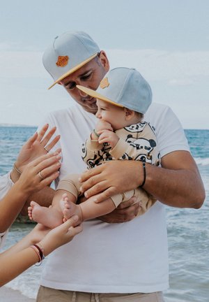 Lichtblauwe petten met een gele rand en een bruine logo, kind in een gemêleerd beige outfit, handen reiken ernaar tegen een strandachtergrond.
