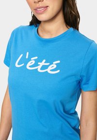 T-shirt en coton bleu avec le texte en cursive blanc "l'été" sur le devant. Col rond et manches courtes, avec une texture douce.