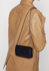 Borsa a spalla in pelle nera con motivo in rilievo del logo e tracolla in catena argentata indossata sopra un giubbotto di pelle beige.